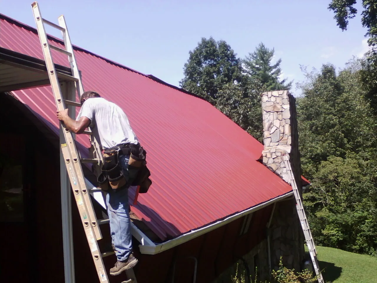 Roofer on a ladder beside a red metal roof and stone chimney — Soffit & Fascia Repair by Roofing Brothers in Mount Sherman